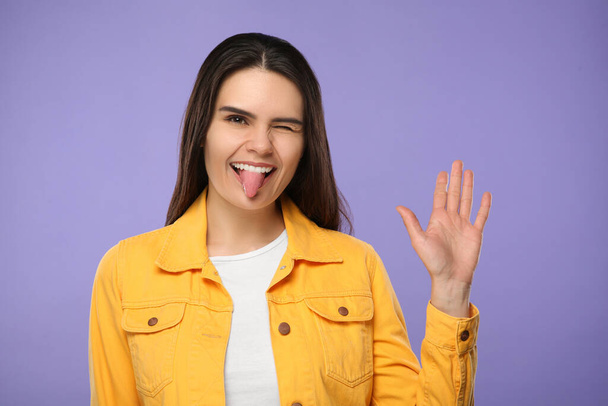 Šťastná mladá žena ukazuje svůj jazyk na fialovém pozadí - Fotografie, Obrázek