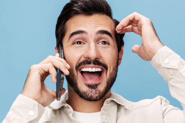 Großaufnahme Porträt eines brünetten Mannes am Telefon, Lächeln mit offenem Mund Glück, auf blauem Hintergrund in weißem T-Shirt und Jeans, Kopierraum. Hochwertiges Foto - Foto, Bild