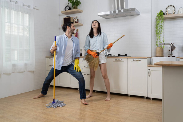 Joyful woman singing songs and dancing with closed eyes while cleaning kitchen at home. Happy young couple having fun while doing cleaning the floor at home and pretend to sing song with mop. - Фото, изображение