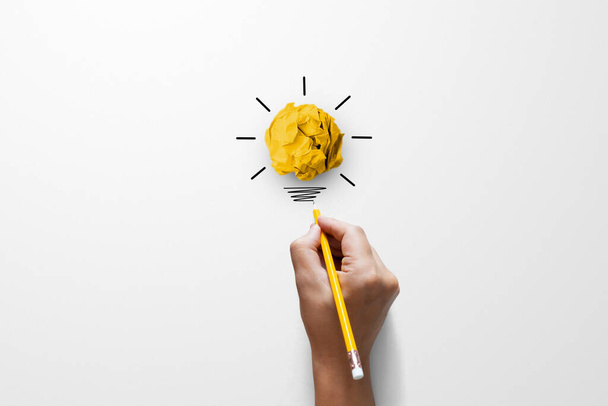 Ideas de pensamiento creativo y concepto de innovación. Pelota de chatarra de papel color amarillo con símbolo de bombilla sobre fondo blanco y mano sosteniendo lápiz amarillo - Foto, imagen