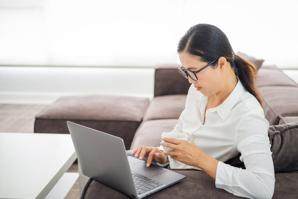 adult woman doing research work for her business. Asian woman sitting on sofa relaxing while browsing online shopping website. Happy lady browsing through the internet during free time at home. - Photo, Image