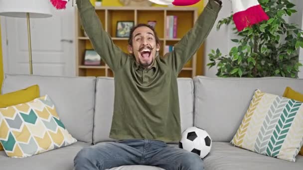 Joven hombre hispano apoyando equipo de fútbol sosteniendo bufanda en casa - Imágenes, Vídeo