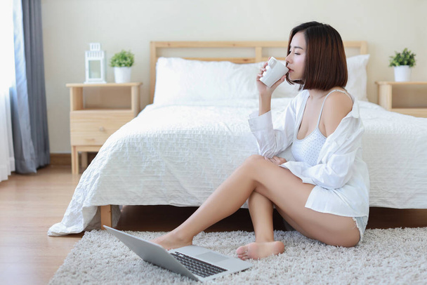 Mulher asiática feliz em casual sentado no chão e segurando xícara de café para o café da manhã enquanto usa o computador em casa, mulher de negócios de beleza com fundo do quarto. Conceito de tecnologia de estilo de vida - Foto, Imagem