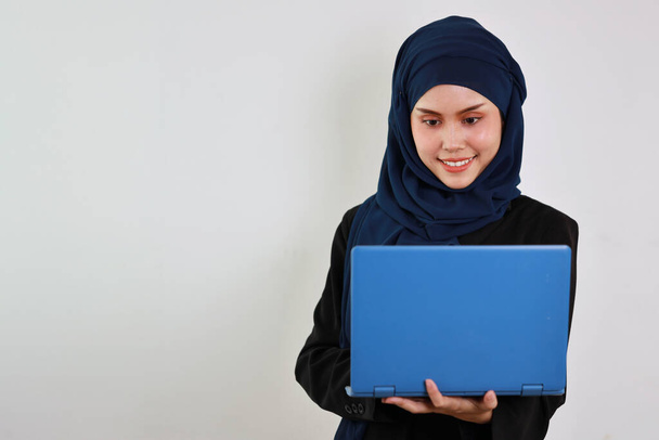 Szczęśliwy inteligentny azjatycki biznes kobieta w sukience muzułmańskiej stoi podczas korzystania z komputera i uśmiecha się z ufnością w studio. Odizolowany biały portret tła z piękną twarzą w hidżabie. - Zdjęcie, obraz