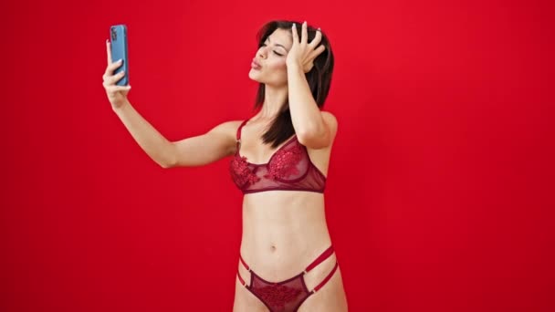 Νεαρή καυκάσια γυναίκα φορώντας εσώρουχα κάνουν selfie από το smartphone σε απομονωμένο κόκκινο φόντο - Πλάνα, βίντεο