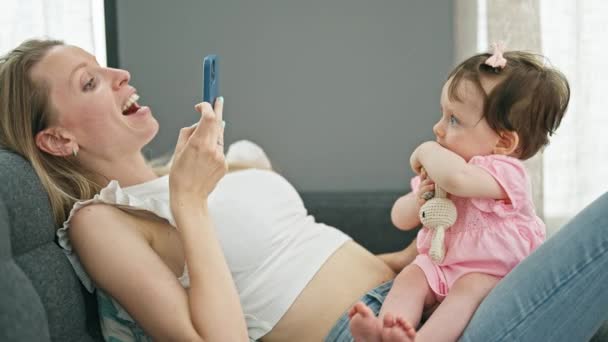Mother and daughter sitting on sofa recording video to baby at home - Footage, Video