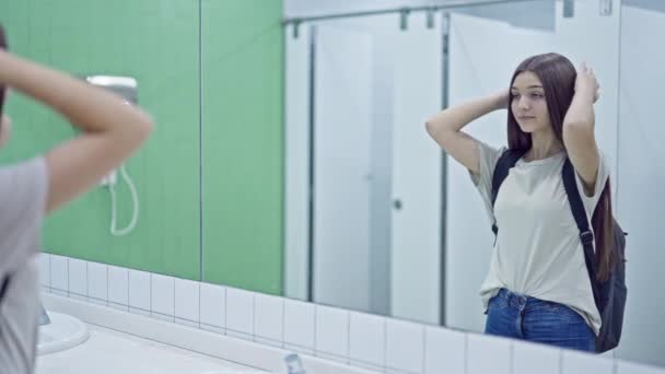 Молодая красивая студентка смотрит на зеркало расчесывая волосы в ванной комнате - Кадры, видео