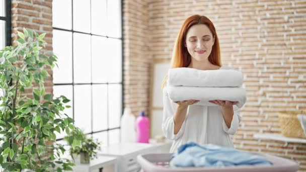 Молодая рыжая женщина улыбается уверенно держа сложенные полотенца в прачечной - Кадры, видео