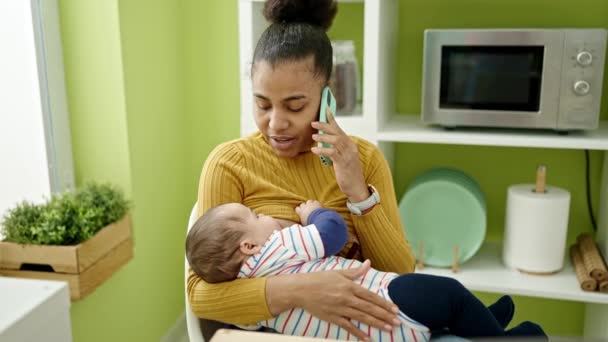 Madre e hijo hablando en el teléfono inteligente amamantando al bebé en el comedor - Metraje, vídeo