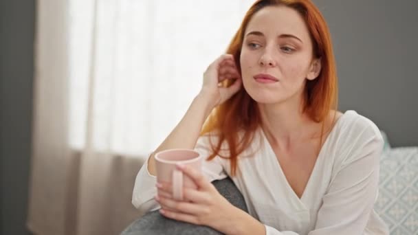 Fiatal vörös hajú nő kávézik otthon a kanapén ülve - Felvétel, videó