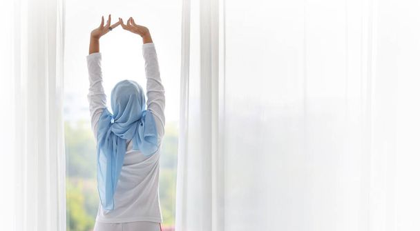 Rückseite schöne asiatische Frau trägt weiße muslimische Nachtwäsche und streckt ihre Arme aus, nachdem sie morgens bei Sonnenaufgang aufgestanden ist. Nettes junges Mädchen mit blauem Hijab steht und entspannt beim Wegschauen - Foto, Bild