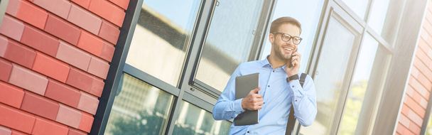 Junger fröhlicher kaukasischer Geschäftsmann mit blauem Hemd und Brille hält seinen Laptop in der Hand, telefoniert und lächelt, während er draußen spaziert. Geschäftsleute-Konzept unter Einsatz moderner Technologien - Foto, Bild