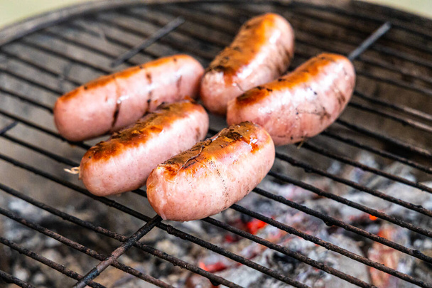 The Group of delicious pink sausages on a black metal grille fried for a picnic are in a fireplace in the forest - Photo, Image
