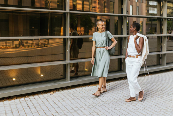 örömteli interakció két üzletasszony, az egyik afro-amerikai, a másik kaukázusi között, ahogy sétálnak az utcán. Mosolyognak és boldog pillantásokat cserélnek, pozitív fényt sugároznak. A fénykép. - Fotó, kép