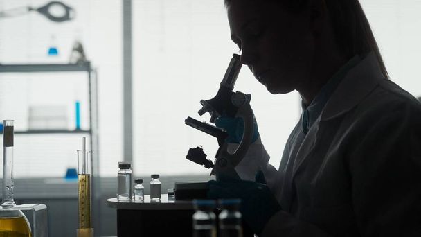 Medical scientific laboratory. Side view of a dark silhouette of a female scientist looking under a microscope, doing an analysis of a test sample close up. Ambitious biotechnology specialist, working - Zdjęcie, obraz