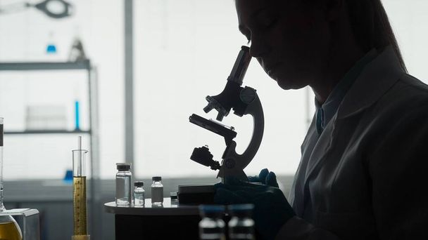 Medical scientific laboratory. Side view of a dark silhouette of a female scientist looking under a microscope, doing an analysis of a test sample close up. Ambitious biotechnology specialist, working - Zdjęcie, obraz