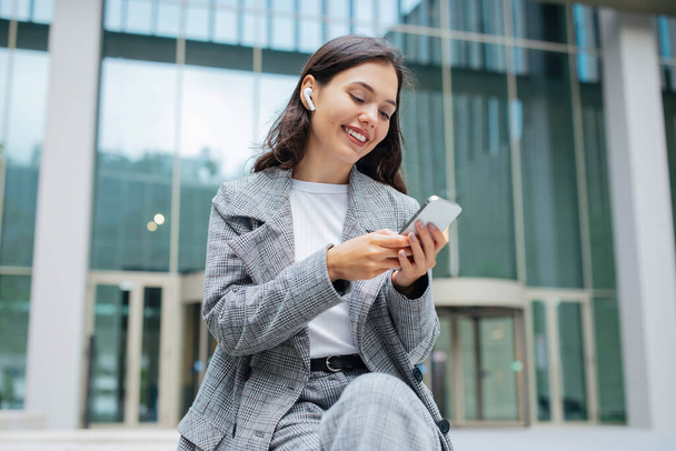 Happy Young Woman In Suit Korzystanie z telefonu komórkowego z bezprzewodowymi słuchawkami Słuchanie muzyki lub biznesu Podcast Online, Siedząc w pobliżu budynku biurowego w obszarze miejskim na zewnątrz. Nowoczesne miasto i Gadżet Styl życia - Zdjęcie, obraz