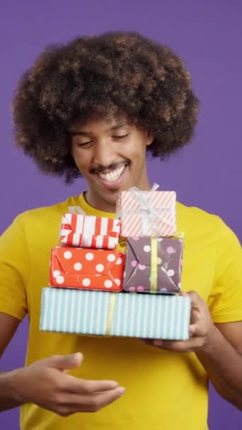 Hombre africano sonriente con el pelo rizado sosteniendo muchos regalos mientras mira la cámara en el estudio - Imágenes, Vídeo
