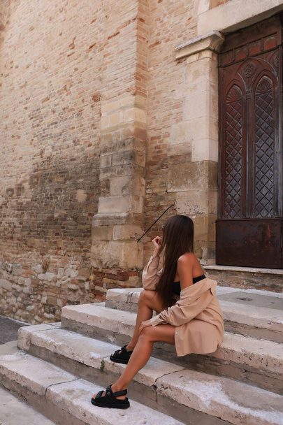 Una giovane ragazza bruna dai capelli fluenti in abito beige per le strade dei vicoli dItalia al tramonto con vasi di fiori alle pareti. Foto de alta calidad - Foto, Imagen