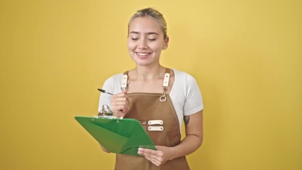 Jeune belle femme hispanique portant tablier écriture sur presse-papiers sur fond jaune isolé - Séquence, vidéo
