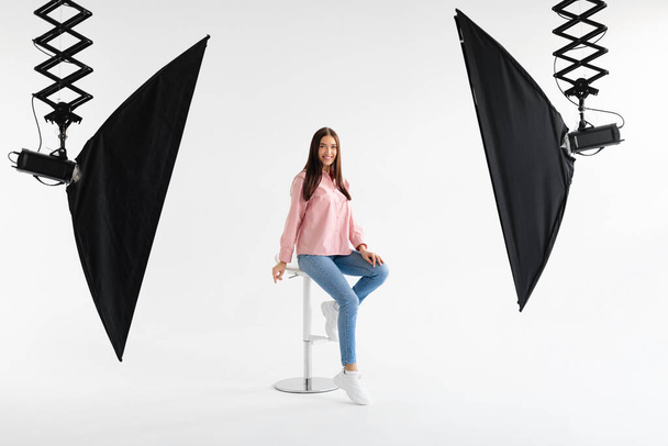 Счастливая европейская дама-модель в повседневной одежде, сидящая на стуле и позирующая на белом фоне в фотоателье, улыбающаяся на камеру, снимающая на телефон и делающая контент для магазина, в полный рост - Фото, изображение