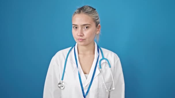 Νεαρή όμορφη ισπανίδα γιατρός στέκεται με σοβαρή έκφραση λέγοντας όχι με το δάχτυλο πάνω από απομονωμένο μπλε φόντο - Πλάνα, βίντεο