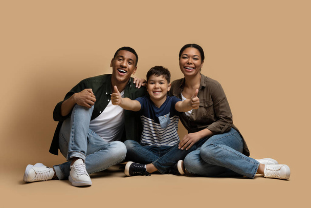 Cheerful happy black family father, mother and school aged son wearing casual outfits posing together on beige background, parents and kid sitting on floor, showing thumb ups and smiling at camera - Photo, Image