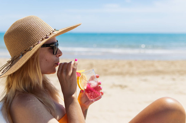 Una rubia sexy en bikini brillante está tomando el sol en una tumbona con un cóctel en la mano junto al mar. Una hermosa chica está disfrutando de sus vacaciones de verano en un complejo caro, Foto de alta calidad - Foto, Imagen