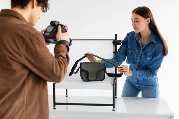Команда фотографа-мужчины и менеджера smm делает фотографии стильной сумочки на белом фоне платформы в современной фотостудии, женщина помогает делать контент фотосессии - Фото, изображение