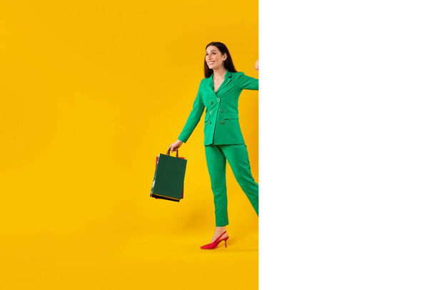 Σχεδιασμός διαφήμισης για ψώνια. Ευτυχισμένο κομψό γυναίκα με τσάντες shopper κρυφοκοιτάζω από λευκό λευκό πανό με χώρο για διαφήμιση, κίτρινο φόντο στούντιο, mockup. Πλήρες μήκος - Φωτογραφία, εικόνα