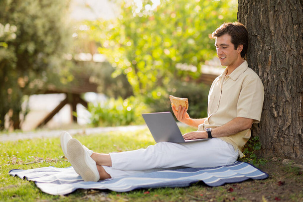 Szczęśliwy przystojny biały millenium mężczyzna niezależny wykonawca pracujący w publicznym parku, facet siedzi na kocu pod drzewem, jedzenie kanapki, pisanie na klawiaturze laptopa, praca online, przestrzeń kopiowania - Zdjęcie, obraz