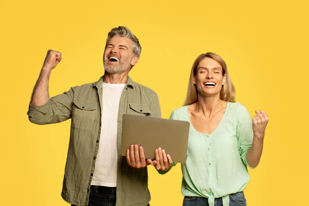 Overjoyed middle aged couple shaking fists holding laptop computer and gesturing yes, celebrating great news on yellow background, studio shot - Foto, Imagem