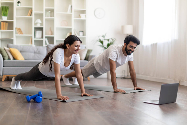Sporty young indian couple in sportswear planking together in living room, happy eastern spouses having online fitness class or video call with trainer from home, using laptop computer, copy space - 写真・画像