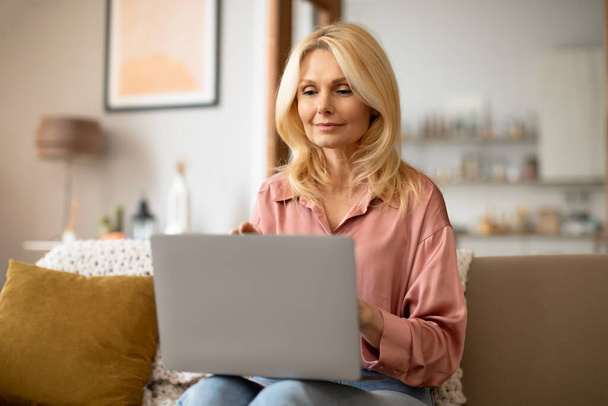 Tecnología, gente de la Edad Media. Retrato de la señora madura sosteniendo ordenador portátil en el regazo de trabajo desde casa en línea, navegar por Internet sentado en cómodo sofá en el interior de la sala de estar - Foto, Imagen
