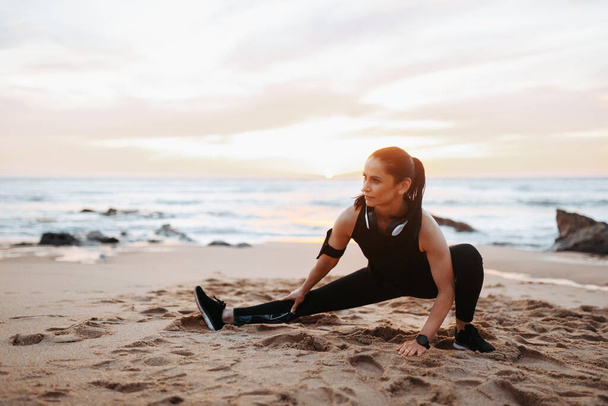 Serious senior caucasian woman in sportswear and headphones with phone doing stretching for legs on beach in morning, outdoor. Sports, workout and active lifestyle, body care - Foto, immagini