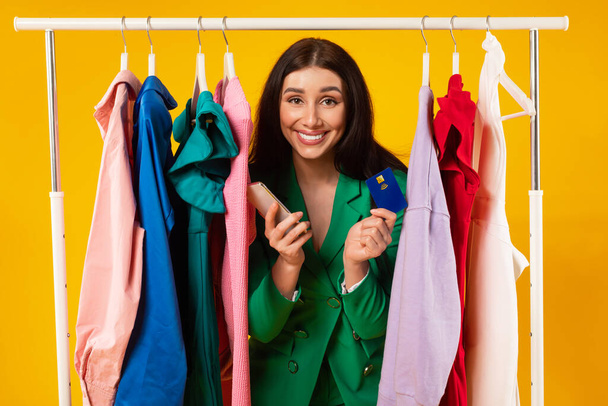 Cheerful young shopaholic lady buying clothes online, woman customer holding cellphone and credit card, posing near clothing rail over yellow studio background - Φωτογραφία, εικόνα