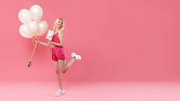 Full length of happy woman celebrating birthday holding bunch of balloons and present box with gift, standing on pink background, panorama with free space, place for advertisement - Photo, image