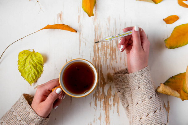 Top view of female hands holding cup of tea and thermometer on shabby table with yellow scattered leaves , autumn composition - Fotoğraf, Görsel