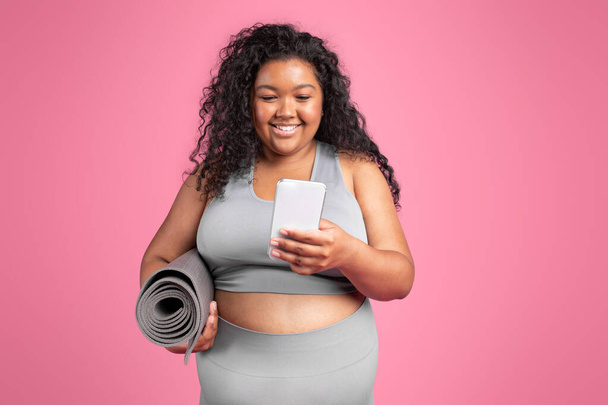 Hymyilevä musta pullea nainen urheiluvaatteet matto chattailuun älypuhelimeen, seisoo vaaleanpunainen studio tausta. Urheilu app, blogi laihtuminen ja kehon hoito, kouluttaja oppitunti etänä - Valokuva, kuva