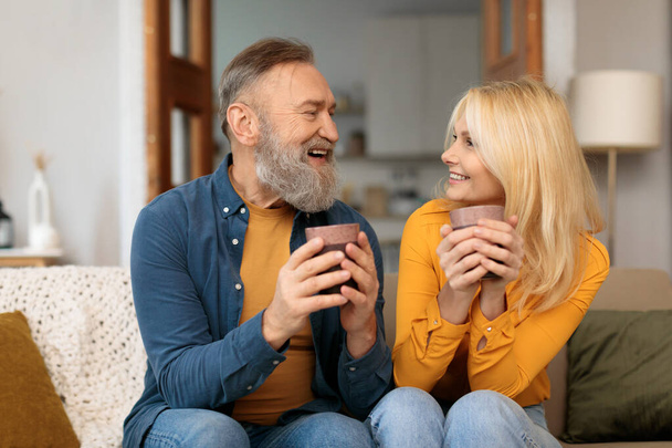 Houden van senior Europese paar bonding terwijl het drinken van koffie in de woonkamer, gelukkig volwassen man en vrouw vasthouden van bekers met warme drankjes en genieten van weekend, tijd samen doorbrengen thuis - Foto, afbeelding