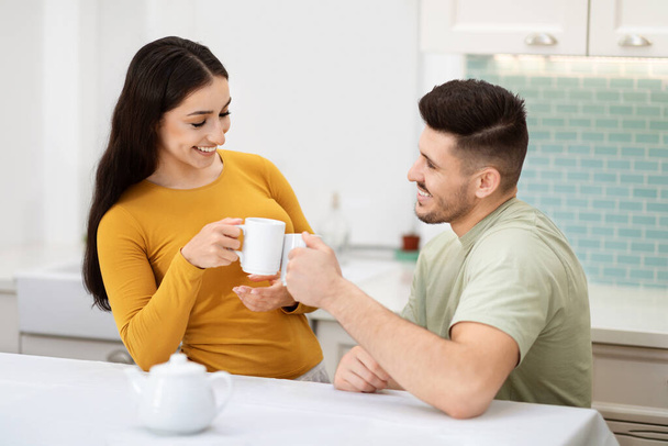 Kaunis nuori mies ja nainen juomassa teetä yhdessä kotona, onnellinen rakastava tuhatvuotinen pari istuu keittiössä, hurraavat valkoiset kahvimukit ja hymyillen. Rakkaus, kiintymys, avioliitto, suhteet - Valokuva, kuva