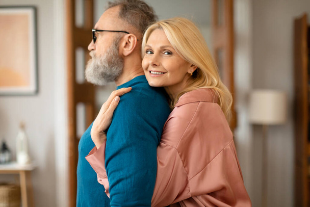 Élvezzük a reggelt együtt. Boldog szerető idős nő ölelkező szakállas férj hátulról állva otthon kényelmes belső, pózol a modern nappaliban, és mosolygós a kamera. Szerelmi kapcsolat - Fotó, kép