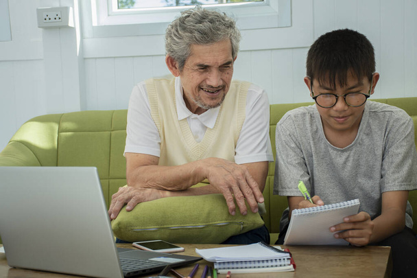 Starszy dziadek pomaga wnukowi odrobić pracę domową przy laptopie w salonie, udzielając wyjaśnień i instrukcji, jak pracować krok po kroku.. - Zdjęcie, obraz