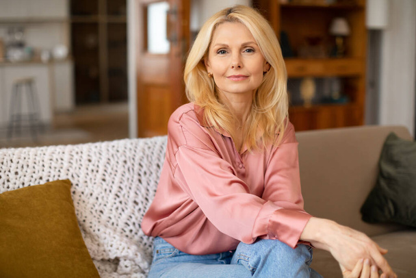 Relaxed Mature Woman Posing On Couch In Cozy Home Interior, Smiling At Camera. Blonde Lady In Casual Enjoying Comfort Of House On Weekend, Sitting And Resting On Sofa In Living Room - Foto, Imagen
