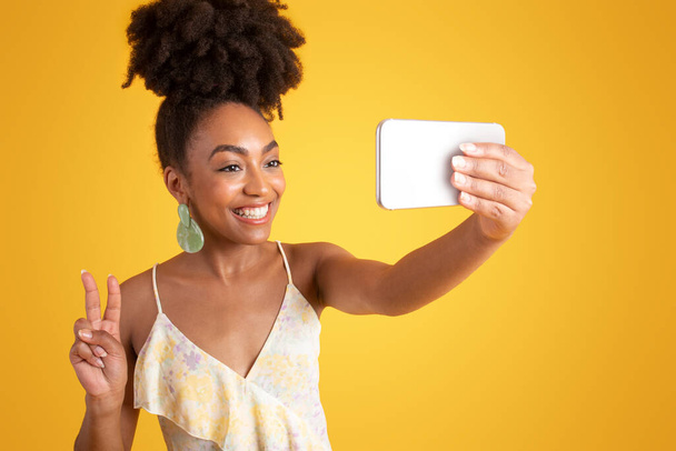 Веселая молодая африканская американка в платье делает селфи на смартфоне, делает знак мира, изолированная на желтом фоне студии. Стиль жизни, модный блог, реклама и предложение, гаджет для фото - Фото, изображение