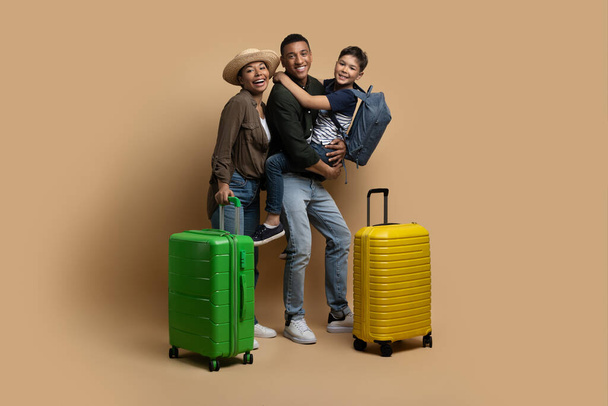 Concept de voyage en famille. Des parents afro-américains excités voyageant avec leur fils préadolescent, posant avec des valises à bagages et des sacs à dos sur fond beige en studio, espace vide - Photo, image