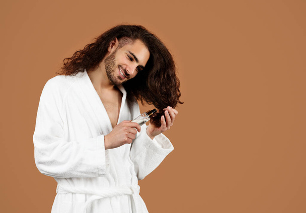 Male Haircare. Happy Brunette Young Man Applying Serum On Long Curly Hair For Repair, Posing Wearing White Bathrobe Standing Over Brown Studio Background. Split Ends Treatment - Fotoğraf, Görsel