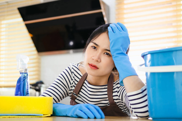 Die schöne asiatische Hausfrau fühlt sich nach der Hausarbeit müde. Erschöpfte Haushälterin in brauner Schürze und blauen Gummihandschuhen sitzt in Küche mit Hausreinigungsprodukten. - Foto, Bild