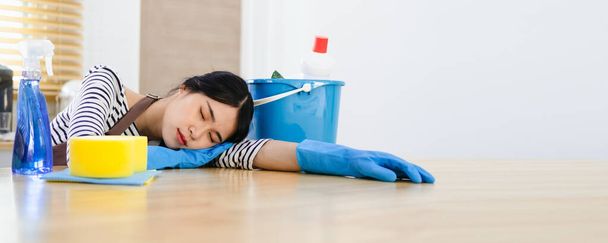 Beautiful Asian housewife feeling tired after doing housework. Exhausted housekeeper in brown apron and blue rubber gloves sitting in kitchen with home cleaning products. - Foto, Imagem