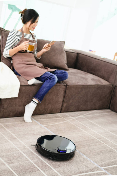 Robot vacuum cleaner cleaning the living room. Young woman enjoy rest, sitting on sofa at home - Foto, afbeelding